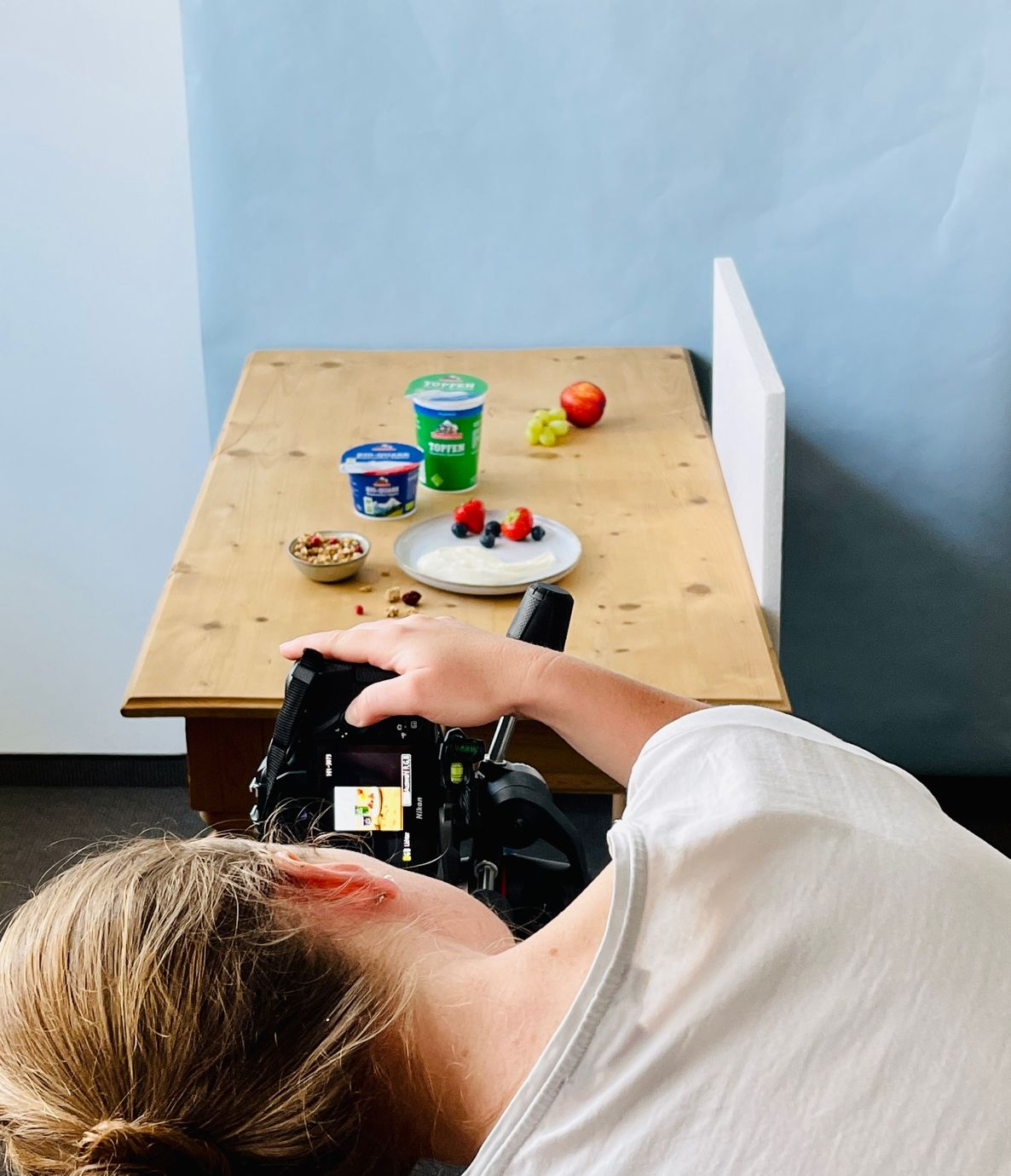 Produktfotografie-Setup mit Kamera aus flacher Perspektive: Bergbauernmilch-Joghurtbecher, Beeren und Müsli auf einem Holztisch vor hellblauem Hintergrund.